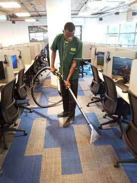 A Bitutam cleaner in green uniform vacuuming an office floor